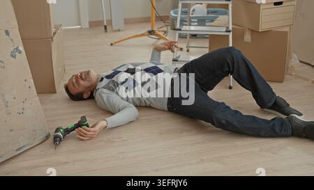 Man lying unconscious on floor with hand near cordless drill beside stacked cardboard boxes and ladder in building interior new home moving scene; acc Stock Photo