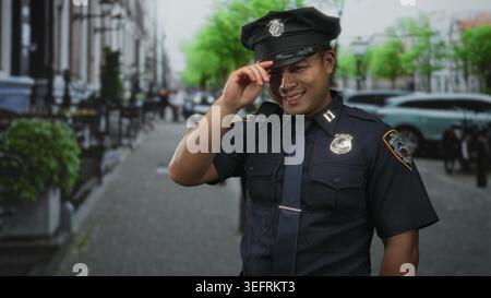 Young chinese policeman tipping hat with smiling gesture on urban street lined with parked cars; pride duty. Stock Photo