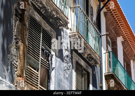 Funchal, Madeira, May 11th 2026. Building facades under a blue sky. One building has peeling dark paint exposed stone and damaged shutters. Stock Photo