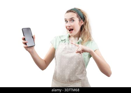 A surprised housewife cook woman wearing an cooking apron holds a smartphone and pointing at it. Isolated on a white background, for food apps or cook Stock Photo