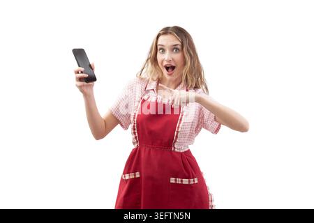 A surprised housewife cook woman wearing an cooking red apron holds a smartphone and pointing at it. Isolated on a white background, for food apps or Stock Photo