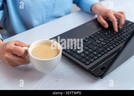 Woman working on laptop while holding coffee cup in modern office workspace, illustrating productive remote work and comfortable digital lifestyle Stock Photo