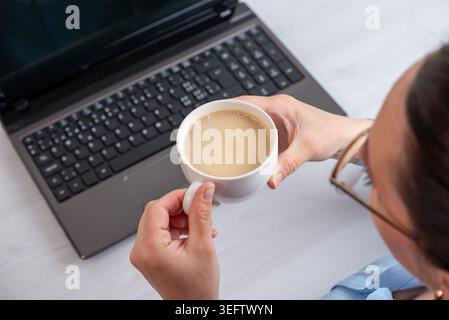 Woman holding coffee above laptop keyboard in relaxed workspace setting, capturing modern freelance lifestyle and remote office atmosphere Stock Photo