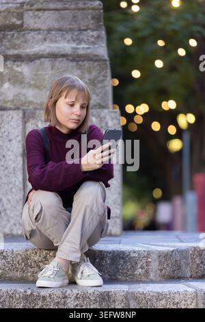 Woman sitting on stone steps at plaza by stone monument holding phone with backpack straps visible Stock Photo