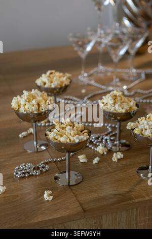 Popcorn is served in glass bowls on a wooden table set for a cinema party with silver decor and party drinks. The scene is ready for guests to have fu Stock Photo