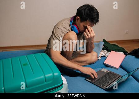 Young man feeling stressed while contemplating his travel plans, sitting with a laptop, open suitcase, and clothes, preparing for a trip or remote Stock Photo