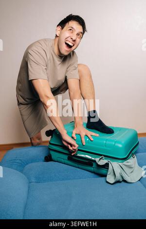 Vertical portrait of angry traveler male struggling to zip overpacked green suitcase for a trip, frowning and making a funny face, experiencing Stock Photo