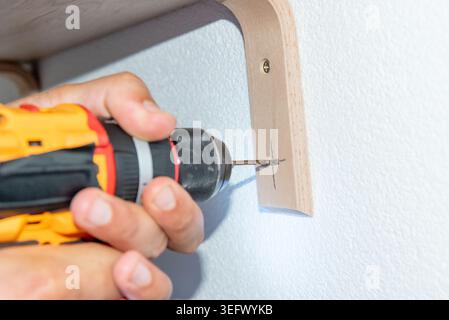 Man Builder Carpenter hangs wooden shelf on brackets on wall using Cordless hammer Drill Stock Photo