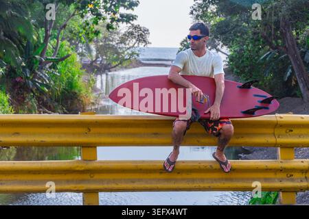 Adult male surfer sitting relaxed on a yellow road bridge railing, holding a red surfboard and looking to the side, tropical river and ocean visible i Stock Photo