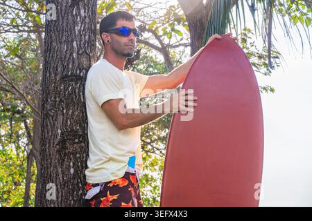 Athletic adult man in t-shirt and boardshorts leaning against a tree trunk holding a red surfboard vertically, dappled tropical light, trees and sky i Stock Photo