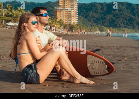 Stylish young couple sitting close together on the beach at golden hour, sharing a tender and loving moment while enjoying the warm sunset light with Stock Photo
