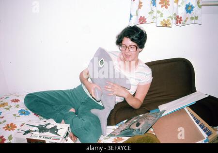 A college student lies on a bed at home in Skokie, Illinois, in July 1976, holding a stuffed shark beside a shark‑themed record album and a Revell 1/48 scale Ju‑87 Stuka model kit. Domestic interior scene typical of mid‑1970s American youth culture. Stock Photo