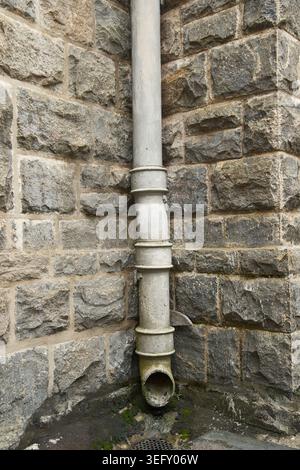 grey metal drainage downpipe base with gutter on stone masonry corner wall, closeup soft focus of ancient plumbing rainwater management Stock Photo