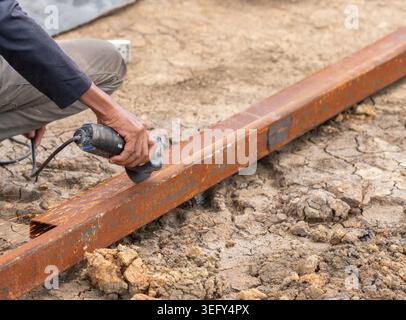 A worker uses a handheld electric angle grinder to smooth and cut a rusty steel beam on an outdoor construction site, creating bright sparks against t Stock Photo
