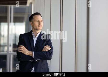 Thoughtful businessman executive standing by office window with folded arms Stock Photo
