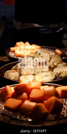 Variety of Chinese dim sum including scallion flower buns, fried mantou, and spring rolls. Displayed on metal trays under warm buffet lighting. Dark b Stock Photo