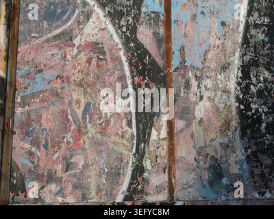 Close-up of a weathered urban wall with peeling paint, rusted strips, and layered graffiti remnants, revealing abstract curves in faded blue, pink, bl Stock Photo