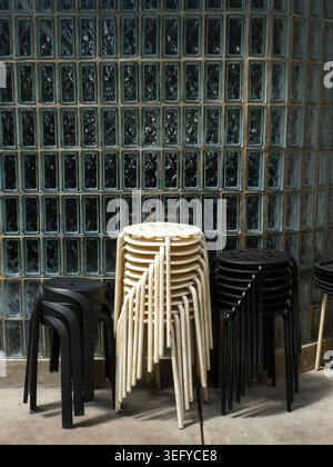 Neatly stacked wooden and black stools sit on a concrete floor against a textured glass block wall, bathed in soft natural light, creating a minimalis Stock Photo