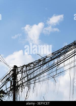Silhouette of a utility pole with tangled powerlines and dangling cables stretching across a bright blue sky with soft white clouds, contrasting chaot Stock Photo