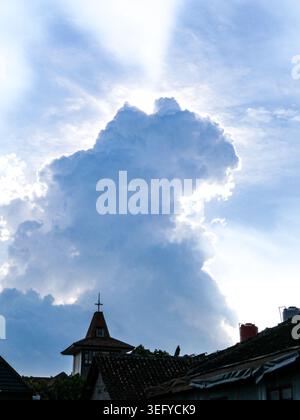 Dramatic backlit cumulus cloud towers above dark tiled rooftops and a small steeple, with radiant sunbeams across a deep blue sky. Quiet urban neighbo Stock Photo