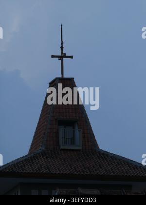 Silhouette of a cross-topped church steeple and terracotta tiled roof against a deep blue twilight sky with a small window detail, evoking serene, sac Stock Photo