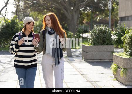 Two female college students walking on campus path, looking at phone, wearing backpacks and beanie Stock Photo