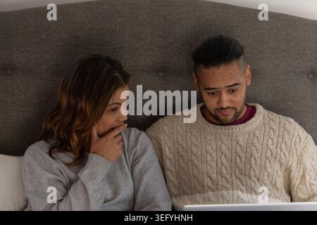 Couple sitting on bed in bedroom, looking at laptop while wearing sweatshirt and cable-knit sweater Stock Photo