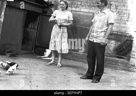 Sloviansk, 1982, courtyard. A young woman in a summer dress holds a tin can as she feeds a flock of pigeons gathered at her feet on the dusty ground. Beside her stands a middle-aged man in a short-sleeved shirt and dark trousers, watching the birds. The background reveals an old brick wall with graffiti, a metal gate, and traditional branch brooms leaning against the structure. This archival photograph preserves the genuine atmosphere of Soviet everyday life, historical nostalgia, and a peaceful Donbas. Stock Photo