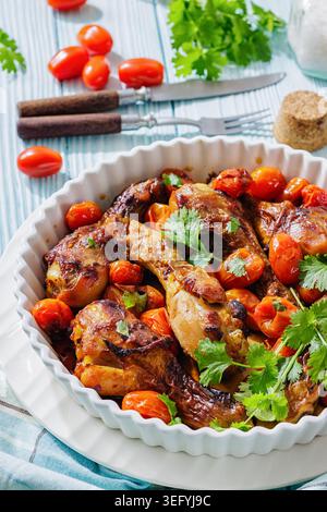 Roasted chicken drumsticks with cherry tomatoes and cilantro in a ceramic baking dish on light blue wooden table with napkin and cutlery, vertical vie Stock Photo