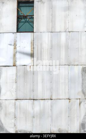 A close-up view of weathered, textured metal siding with a repeating panel pattern. A single window reveals scaffolding and green sheeting behind it. Stock Photo