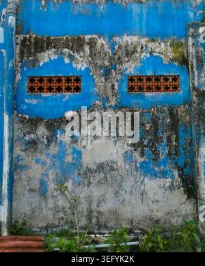 Weathered blue wall with two orange vent grates, peeling plaster over gray concrete, and moss at the base, evoking gritty urban decay and quiet resili Stock Photo