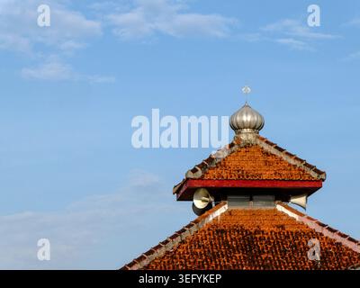 Low angle view of a traditional terracotta orange tiled mosque roof featuring a metallic dome pinnacle against a clear blue sky in Indonesia Stock Photo