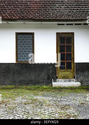 Front view of an old building facade featuring a weathered wooden door and a rectangular window set against a split-tone wall with gravel foreground i Stock Photo