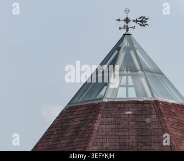 A close-up view of a pointed building roof featuring a glass pyramid structure topped with a decorative metal weathervane against a clear, pale sky Stock Photo