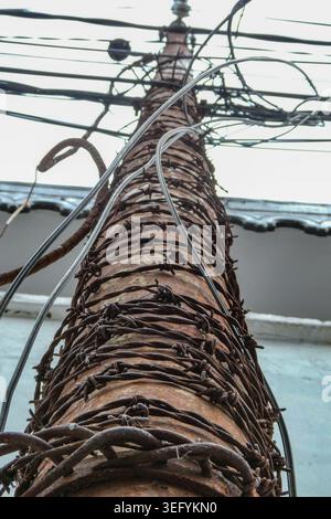 Low angle view of an old rusty utility pole tightly wrapped with sharp barbed wire, featuring tangled overhead electrical cables against a blurred bui Stock Photo
