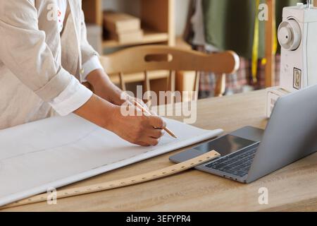 A designer hand sketches garment patterns on paper with a pencil and wooden ruler working on a laptop at a professional fashion studio desk. Stock Photo
