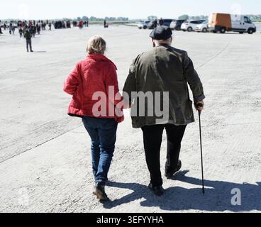 Elderly couple walks hand in hand across a concrete parking lot. The woman wears a bright red jacket and blue jeans, while the man wears a dark coat, Stock Photo