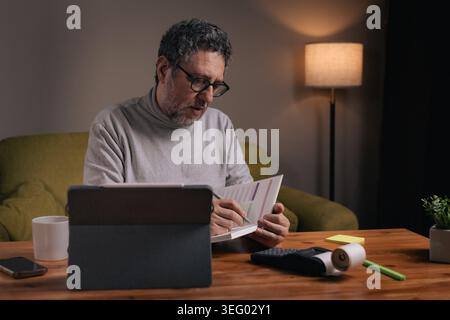 Mature man sitting at home writing financial notes in a planner while using a tablet and calculator. Personal budgeting, expense tracking and financia Stock Photo