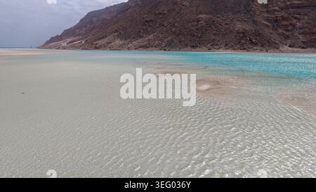 Shallow clear lagoon with rippled sandy seabed, turquoise water and rocky desert mountains under overcast sky, tranquil remote coastline. Stock Photo