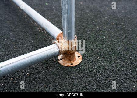 Heavily rusted metal base flange and modular corner clamp securing galvanized steel pipes on a black rubber sport court surface. Stock Photo