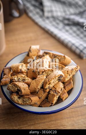 Homemade Italian biscotti cantuccini piled in an enamel bowl and scattered on a rustic wooden table, ideal for coffee or tea breaks. Warm, crunchy golden baked cookies that evoke cozy mornings and casual hospitality. Stock Photo