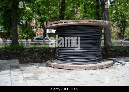 Industrial wooden reel with coiled electrical or fiber optic cable on a stone tile pavement, with a blurred city park and trees in the background. Stock Photo