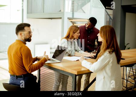 Four people are gathered around a large table in an office space. They are using laptops and discussing ideas. The setting has large windows that let Stock Photo