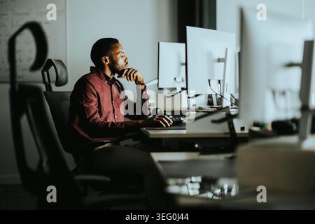 A man in a maroon shirt sits at a desk surrounded by multiple monitors, deeply focused on his work in a contemporary office setting at dusk. Stock Photo
