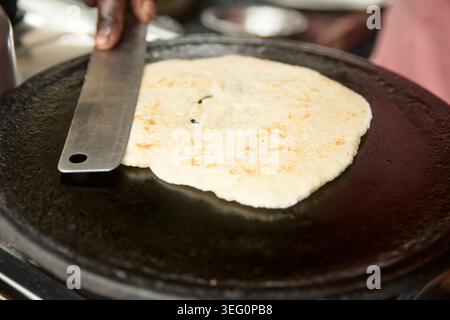 flat metal spatula held by hand pressing roti flatbread, cooking dough on black cast iron griddle pan, closeup soft focus, traditional recipe Stock Photo