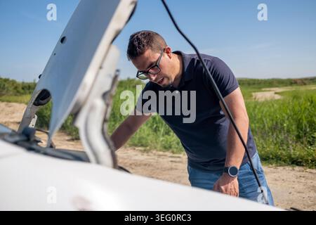 Young man checking under hood of a broken down car, standing on a dirt road in a rural area. Experiencing vehicle trouble Stock Photo