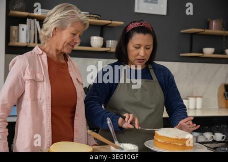 Diverse mature female friends frosting two-layer sponge cake in home kitchen with offset spatula Stock Photo