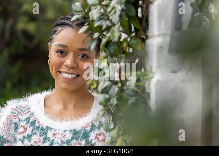 African American woman 20s smiling wearing fuzzy-sweater gold-hoops near white wall with ivy Stock Photo