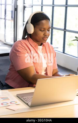 Adult African American woman wearing pink shirt, white headset working at desk with laptop, charts Stock Photo