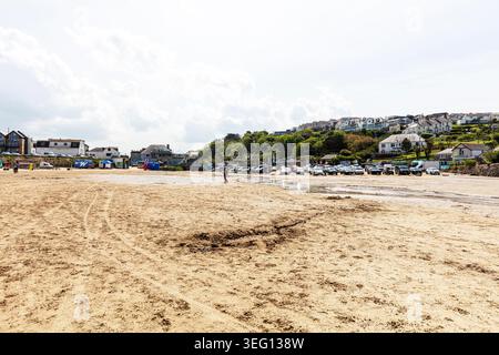 Wide sandy beach at Polzeath in Cornwall with tyre tracks and footprints visible across the sand. A few people walk along the shoreline, while houses Stock Photo